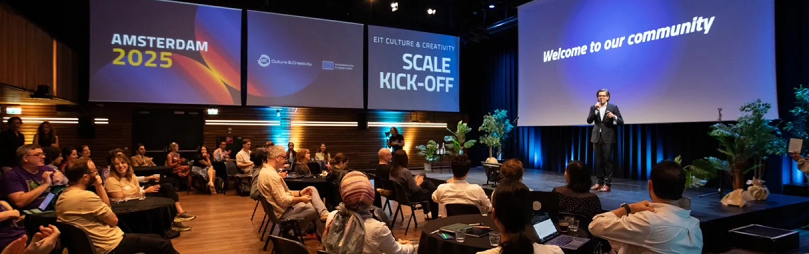 Event participants seated around tables while a speaker on stage welcomes the audience during the “Scale Kick-off” event, with large screens displaying event visuals.