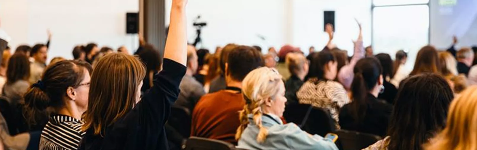 Audience seated in a conference room, with several participants raising their hands to take part in the discussion.