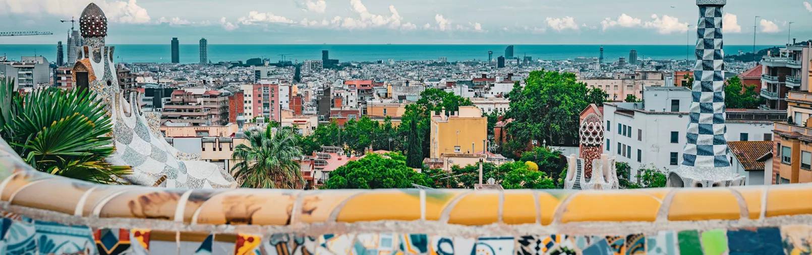 Panoramic view of Barcelona from Park Güell, showing colourful mosaic architecture in the foreground and the city stretching toward the sea.