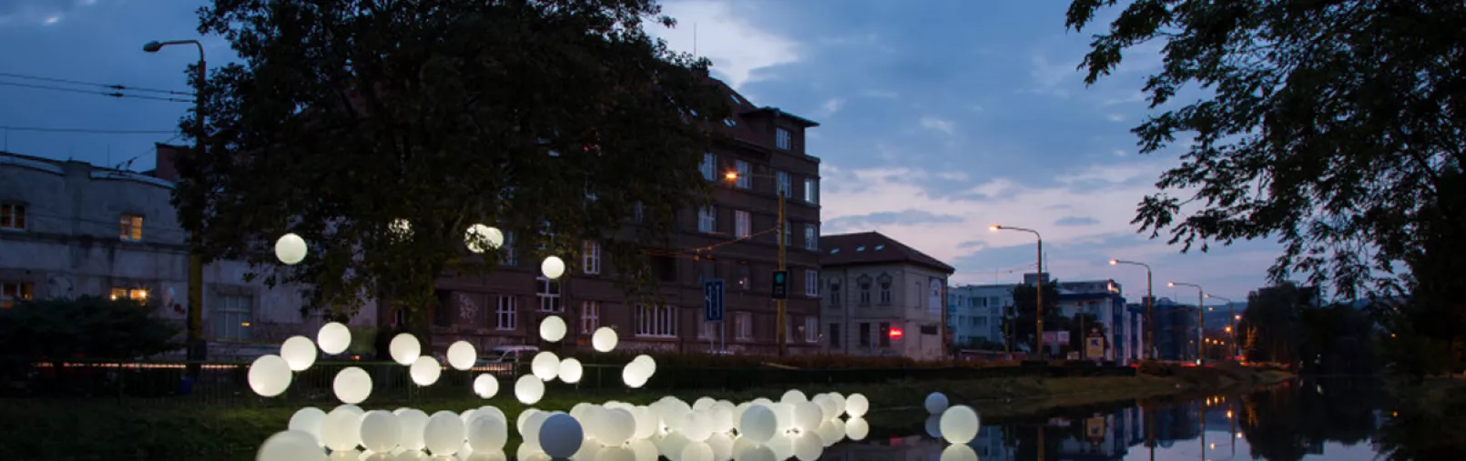 Art installation of glowing white spheres floating on a river at dusk, with surrounding buildings and trees reflected in the water