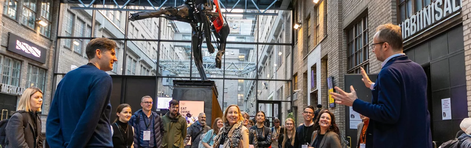 Group of people gathered in an industrial-style event space, listening to a speaker under a large suspended sculpture