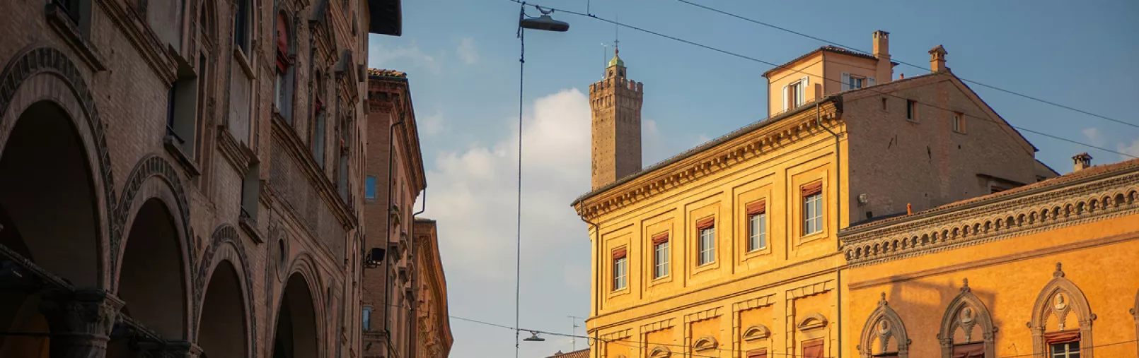 Sunlit historic buildings and porticoes in an Italian city street, with a medieval tower rising in the background under a blue sky