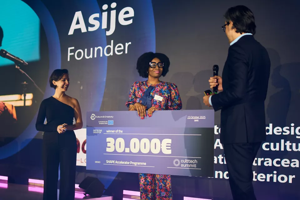 Award ceremony on stage where a winner holds a large cheque for 30,000€, flanked by two presenters during an event.