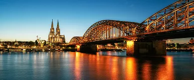 Evening view of Cologne with the illuminated Hohenzollern Bridge spanning the river and the Cologne Cathedral visible in the background.