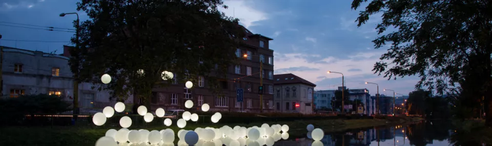 Art installation of glowing white spheres floating on a river at dusk, with surrounding buildings and trees reflected in the water