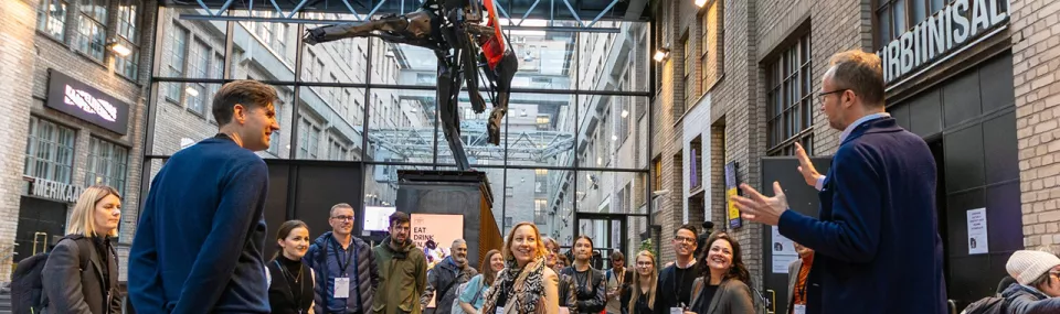 Group of people gathered in an industrial-style event space, listening to a speaker under a large suspended sculpture