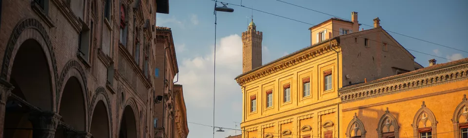 Sunlit historic buildings and porticoes in an Italian city street, with a medieval tower rising in the background under a blue sky